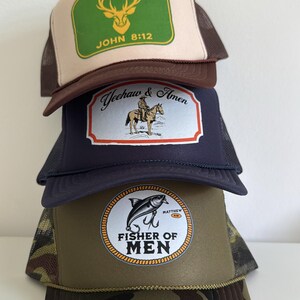Puede incluir: Una pila de cuatro gorras de camionero con diferentes dise&ntilde;os. La gorra superior es beige y marr&oacute;n con un rect&aacute;ngulo verde y el texto "JOHN 8:12". La segunda gorra es azul marino con un gr&aacute;fico de vaquero y caballo. La tercera tiene un gr&aacute;fico de pez y el texto "FISHER OF MEN". La gorra inferior es de camuflaje.