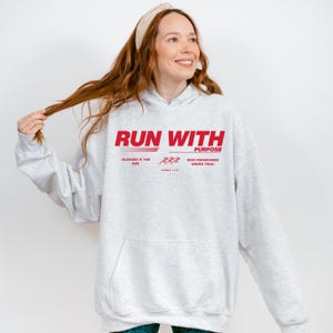 May include: A light gray hoodie with a red graphic that reads "RUN WITH PURPOSE." The text includes the phrases "Blessed is the one" and "Who perseveres under trial." The hoodie has a front pocket and a drawstring hood.