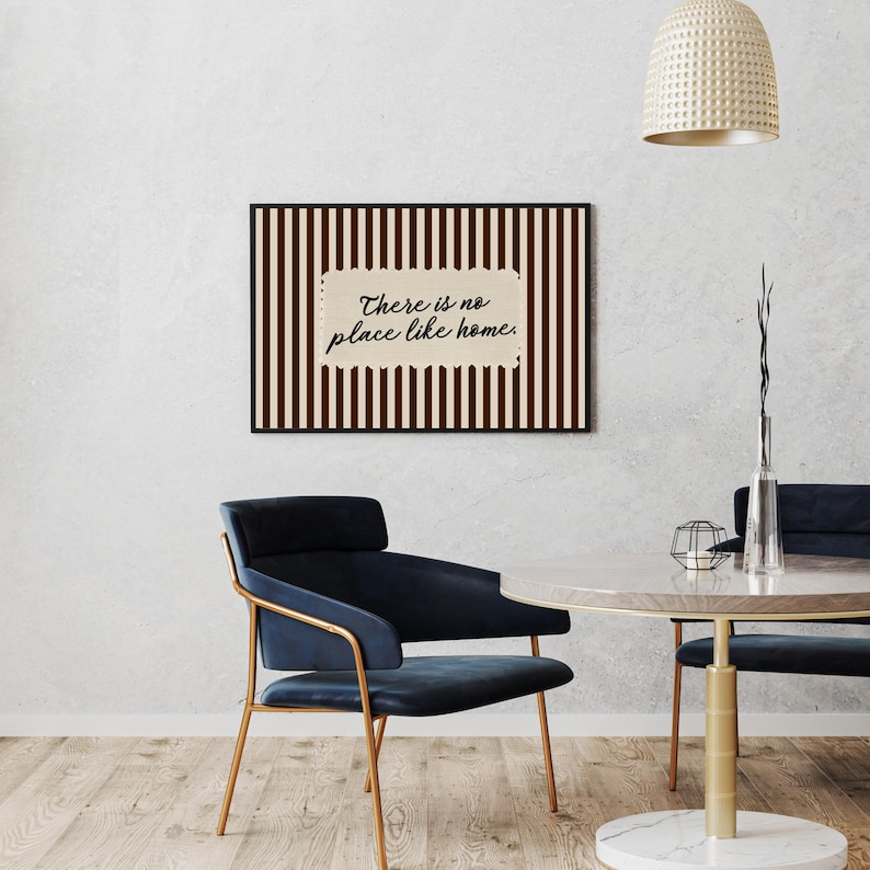 May include: A framed artwork with a black border, displaying a striped pattern in brown and cream. The text "There is no place like home" is written in a cursive font. The image also shows a blue chair and a round table, typical of home decor.