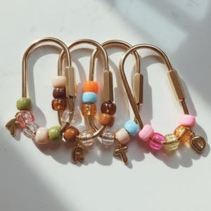 Puede incluir: Llaveros dorados con detalles de cuentas de colores. Cada llavero presenta un diseño curvo con una serie de cuentas en varios colores, incluyendo rosa, verde, naranja y marrón. Los llaveros tienen dijes de letras.