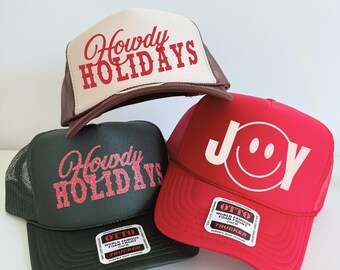 May include: Three trucker hats in the image. One is tan with "Howdy HOLIDAYS" in red script. Another is dark green with the same text in pink glitter. The third is red with "JOY" and a smiley face. All have a mesh back.