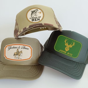 Puede incluir: Tres gorras de camionero con diferentes dise&ntilde;os. Una gorra es de color beige con un patr&oacute;n de camuflaje y un parche circular que dice "FISHER OF MEN". Otra gorra es de color verde oliva con un parche que dice "Yeehaw & Amen". La tercera gorra es de color verde oscuro con un parche verde que dice "JOHN 8:12".