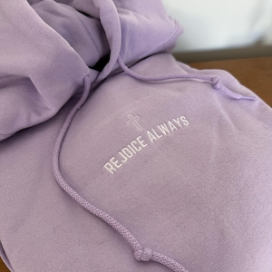 May include: A lavender colored hoodie with a white embroidered cross and the text "Rejoice Always".