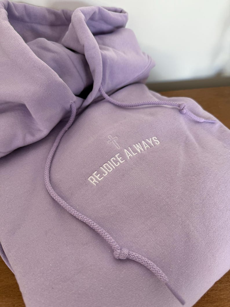 May include: A lavender colored hoodie with a white embroidered cross and the text "Rejoice Always".