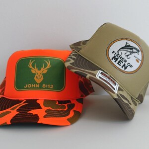 Puede incluir: Dos gorras de camionero. Una es naranja con un rect&aacute;ngulo verde con un ciervo y el texto "JOHN 8:12". La otra es de color canela con un patr&oacute;n de camuflaje y un emblema circular que dice "FISHER OF MEN".