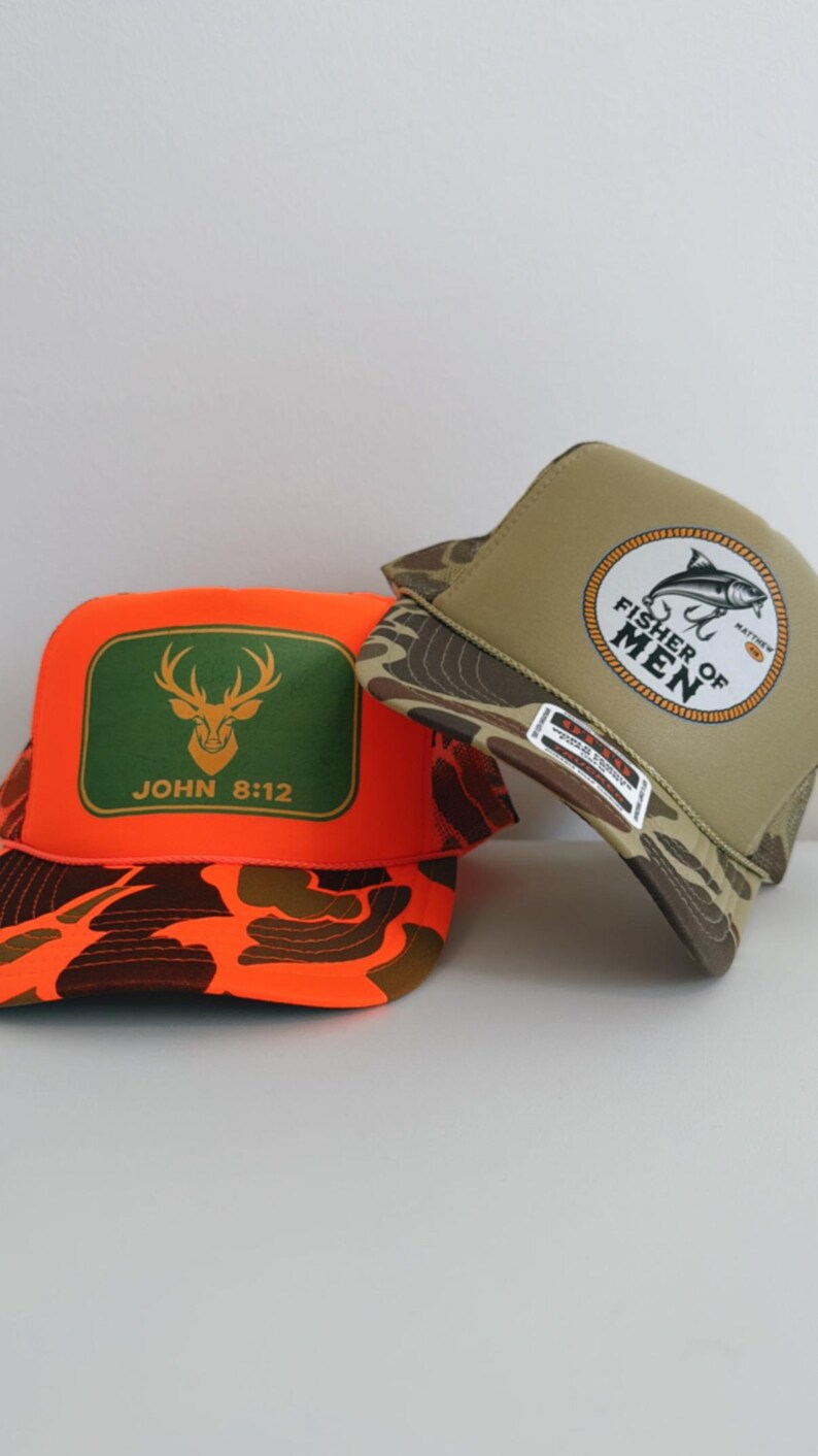 Puede incluir: Dos gorras de camionero. Una es naranja con un rect&aacute;ngulo verde con un ciervo y el texto "JOHN 8:12". La otra es de color canela con un patr&oacute;n de camuflaje y un emblema circular que dice "FISHER OF MEN".