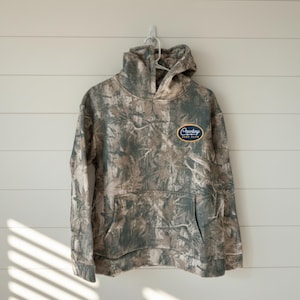 May include: A camouflage-patterned hoodie with a kangaroo pocket and a drawstring hood. The hoodie features an oval patch with the text "Cowboy Surf Club" in a blue and gold design. The garment hangs on a white hanger against a white wall.