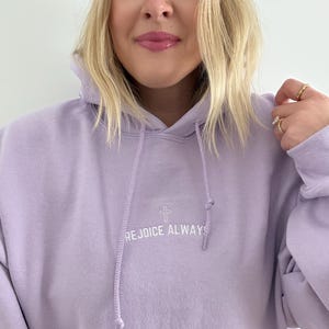 May include: A lavender colored hoodie with a white embroidered cross and the text "REJOICE ALWAYS".