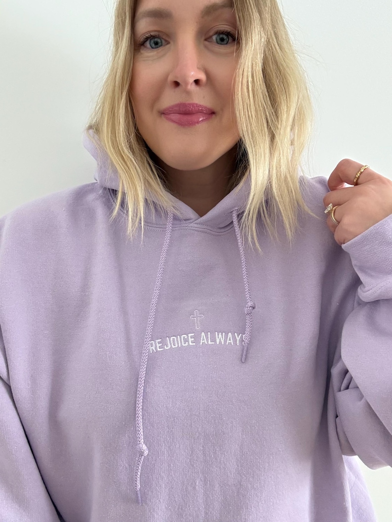 May include: A lavender colored hoodie with a white embroidered cross and the text "REJOICE ALWAYS".