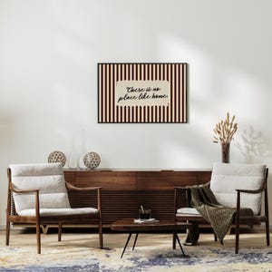 May include: A framed print with the text "There is no place like home" against a striped background. The artwork is above a wooden cabinet and two armchairs with cream cushions. A small coffee table and decorative items complete the interior design.