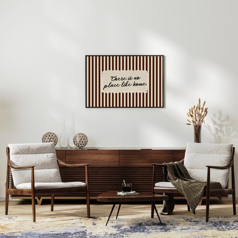 May include: A framed print with the text "There is no place like home" against a striped background. The artwork is above a wooden cabinet and two armchairs with cream cushions. A small coffee table and decorative items complete the interior design.