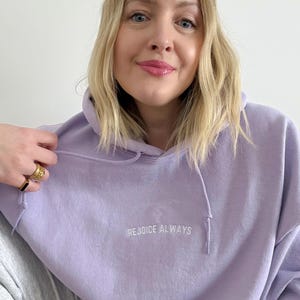 May include: A lavender colored hoodie with a white embroidered cross and the text "REJOICE ALWAYS".