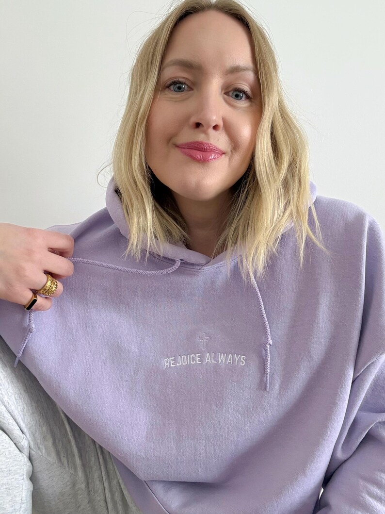 May include: A lavender colored hoodie with a white embroidered cross and the text "REJOICE ALWAYS".
