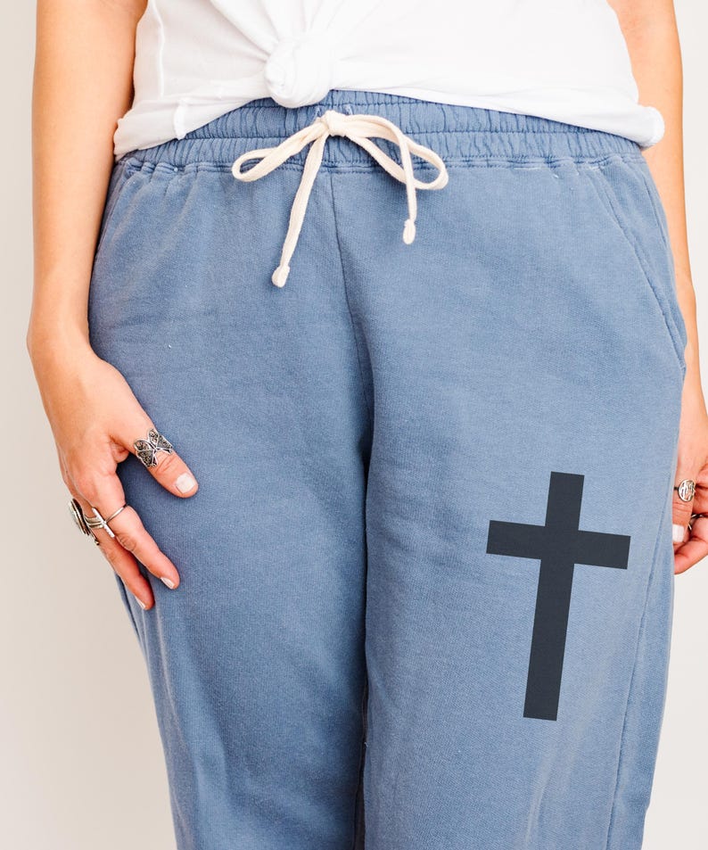 May include: Blue sweatpants with a black cross graphic printed on the right leg. The sweatpants have a drawstring waist.