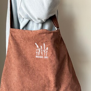 Puede incluir: Una bolsa de mano de pana marr&oacute;n con un dise&ntilde;o bordado blanco de una planta y el texto "IMAGO DEI".