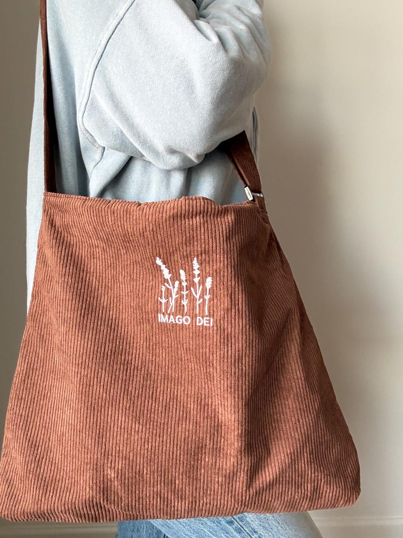 Puede incluir: Una bolsa de mano de pana marr&oacute;n con un dise&ntilde;o bordado blanco de una planta y el texto "IMAGO DEI".