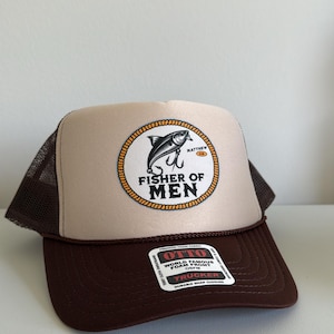 Puede incluir: Gorra de camionero marr&oacute;n y beige con parte trasera de malla. El panel frontal presenta un emblema circular con un dise&ntilde;o de pez y anzuelo, y las palabras "FISHER OF MEN". La gorra tiene una visera curva y cierre a presi&oacute;n.