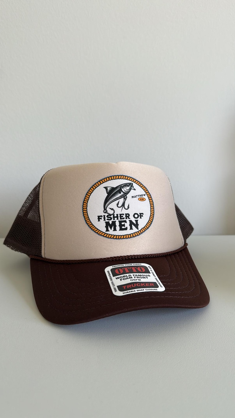 Puede incluir: Gorra de camionero marr&oacute;n y beige con parte trasera de malla. El panel frontal presenta un emblema circular con un dise&ntilde;o de pez y anzuelo, y las palabras "FISHER OF MEN". La gorra tiene una visera curva y cierre a presi&oacute;n.