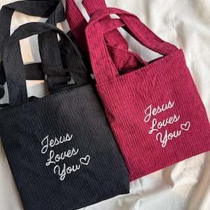 Puede incluir: Dos bolsos tote de pana, uno negro y otro burdeos. Cada bolso tiene una correa larga y el texto bordado "Jesus Loves You" con un símbolo de corazón. Los bolsos están sobre una tela blanca.