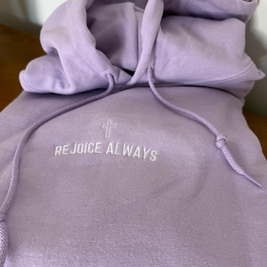 May include: A lavender colored hoodie with a white embroidered cross and the text "REJOICE ALWAYS" on the front.