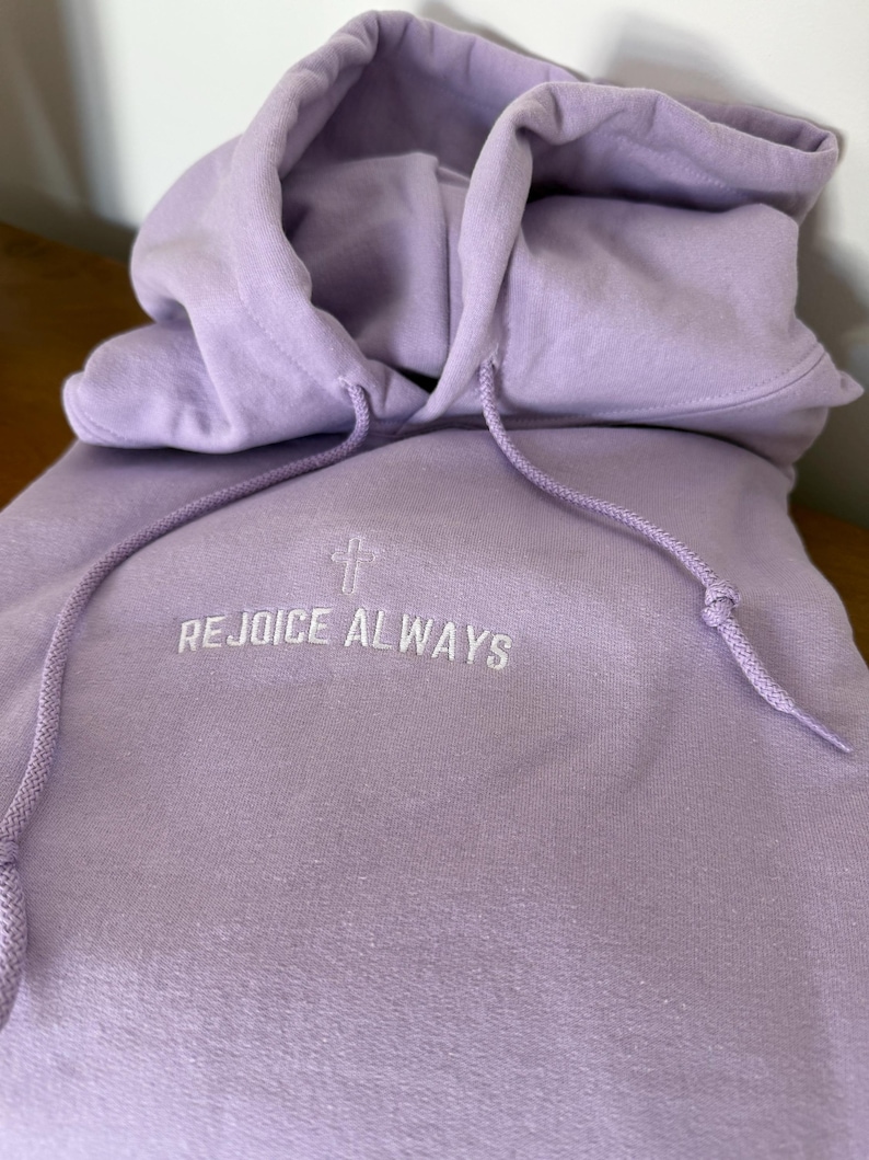 May include: A lavender colored hoodie with a white embroidered cross and the text "REJOICE ALWAYS" on the front.