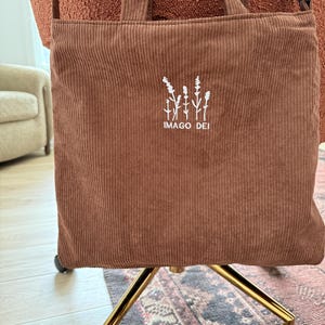 Puede incluir: Una bolsa de mano de pana marr&oacute;n con flores bordadas en blanco y el texto "IMAGO DEI".