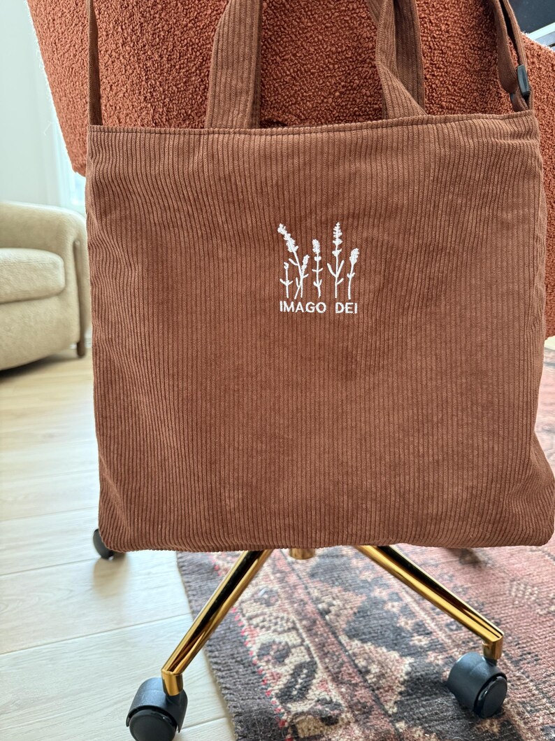 Puede incluir: Una bolsa de mano de pana marr&oacute;n con flores bordadas en blanco y el texto "IMAGO DEI".