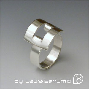 May include: A silver ring with a geometric design. The ring is made of two pieces of metal that are joined together to form a square shape with a cut-out in the centre.