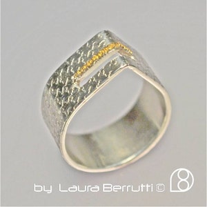 May include: A silver ring with a textured surface and a gold line across the top. The ring is shaped like a rectangle with a cut-out in the middle.