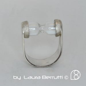 May include: A silver ring featuring a clear hourglass design. The hourglass is set into the band of the ring, which is open on one side.