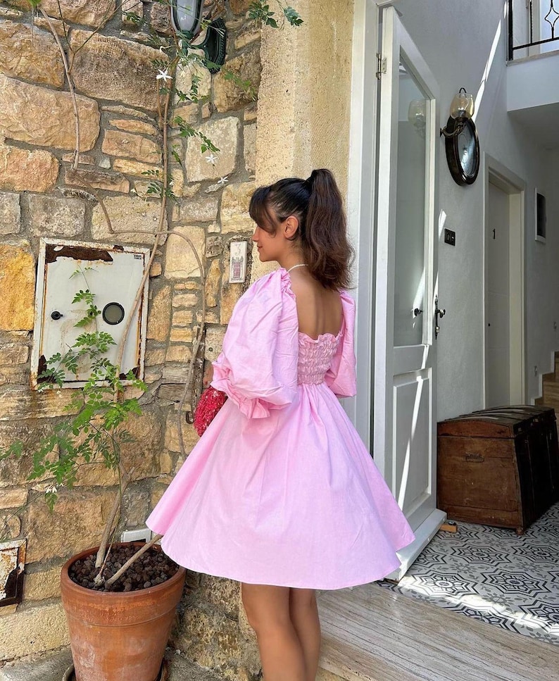 May include: A pink, short-sleeved dress with a square neckline and a gathered waist. The dress has a puffed sleeve design and a flowy skirt.