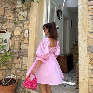 May include: A pink, short-sleeved dress with a square neckline and puffed sleeves. The dress has a gathered waist and a flowy skirt. The model is wearing the dress with a pink sequined handbag.