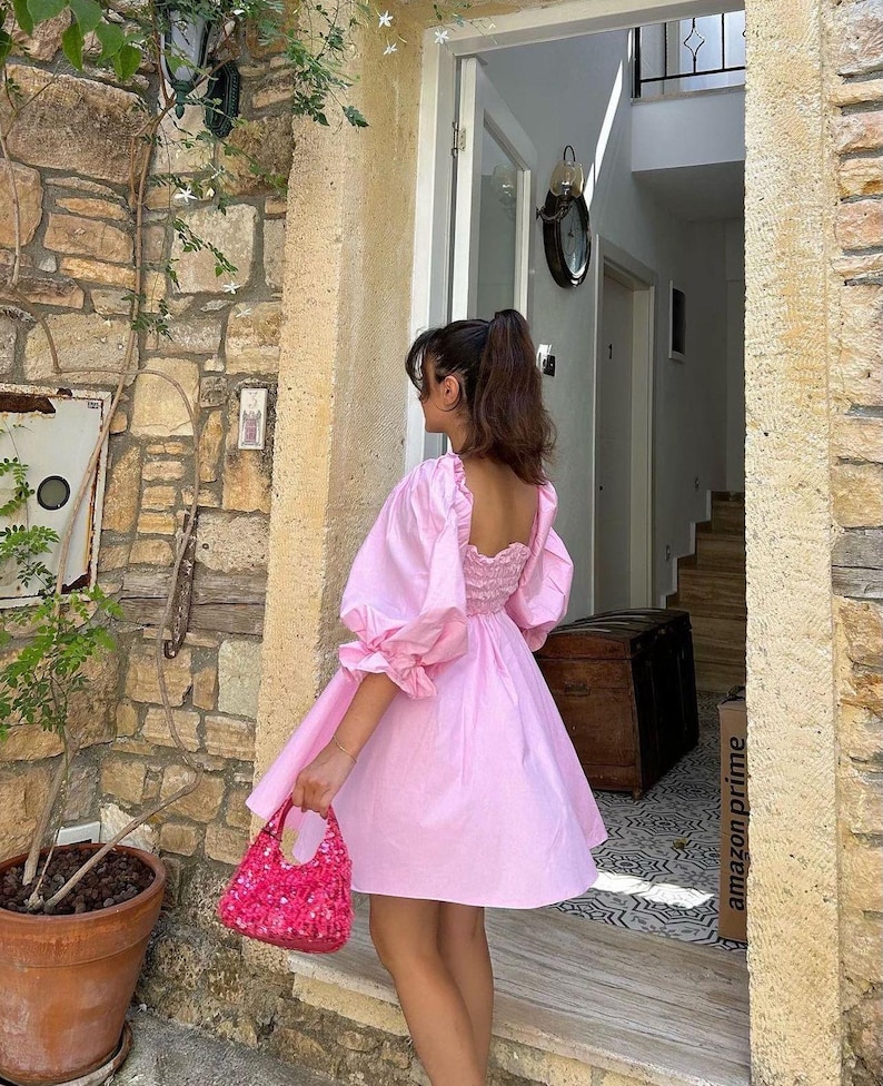 May include: A pink, short-sleeved dress with a square neckline and puffed sleeves. The dress has a gathered waist and a flowy skirt. The model is wearing the dress with a pink sequined handbag.