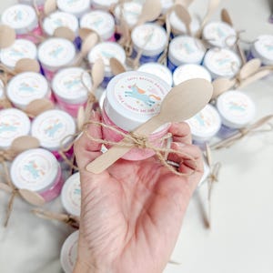 May include: A small clear plastic jar filled with pink substance, topped with a white lid featuring a unicorn graphic and the text Happy Magical Birthday Ada & Florence Are Five. A wooden spoon is tied to the jar with brown twine.