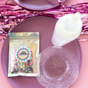 May include: A close-up shot of a purple plate with a clear, decorative bowl, a small wooden spoon, and a packet of colorful confetti. The packet has a label that reads "imagidough FIZZ". A white bottle and pink tinsel are also visible.
