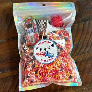 May include: A clear holographic pouch filled with red, yellow, white, and black dyed rice, a small red and white toy race car, a checkered flag on a toothpick, and a red pom-pom. A circular label features the text ImagiDough Grand Prix.