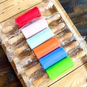 May include: A collection of colorful rolling pins in a wooden tray. The rolling pins have wooden handles and come in red, pink, light blue, orange, blue, and green. Each rolling pin is individually wrapped in clear plastic.