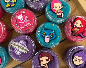May include: A collection of round clear plastic jars filled with colorful slime, each topped with a unique character or logo charm. The slimes are purple, pink, and teal, featuring glitter accents. White lids with the text HUNTRIX are visible.