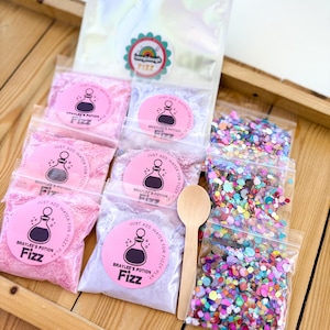 May include: A collection of colorful bath fizz products, including pink, purple, and confetti-like elements, arranged on a wooden tray. The products are packaged in clear plastic bags and feature the text "Braylee's Potion Fizz". A wooden spoon is also included.