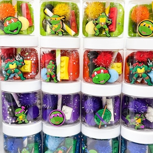 May include: A collection of sixteen clear plastic jars with white lids, each filled with colorful sensory dough, fuzzy pom-poms, small plastic building bricks, and decorative charms featuring Teenage Mutant Ninja Turtles characters.