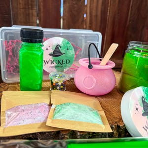May include: A witch-themed potion kit with a green potion bottle, a pink cauldron, and small packets of colorful ingredients. The kit includes a clear container with a "Wicked Potion Kit" label and a jar of green liquid. The items are arranged on a wooden surface.