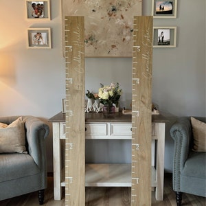 May include: Two wooden growth charts with white painted numbers and names. The charts are mounted on a wall with a white background. The names on the charts are "Cadley Haelin" and "Camille Eaton".