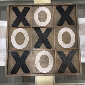 May include: Wooden tic-tac-toe game with a dark brown frame. The game board features alternating black and natural wood "X" and "O" pieces. The game is set on a light brown wooden surface.
