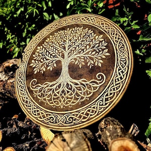 May include: Round wooden wall art featuring a detailed tree of life design. The tree is carved in a light color against a dark brown background. The outer edge has a Celtic knot border. The art is displayed outdoors.