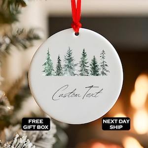 May include: White ceramic Christmas ornament with a watercolor forest scene and the words "Custom Text" in script. The ornament hangs from a red ribbon. The background is blurred, suggesting a festive setting.