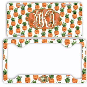 May include: A license plate and frame set featuring a repeating pattern of orange pineapples on a white background. The license plate has a monogram in an orange and green decorative frame.