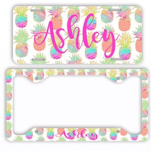 May include: A license plate and frame set featuring a repeating pattern of colorful pineapples on a white background. The name "Ashley" is written in a bright pink script across the license plate.