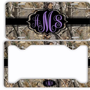 May include: Camouflage-patterned license plate and frame. The plate features a black stripe and a purple monogram within a black decorative frame. The frame has a matching camouflage pattern and a smaller monogram.