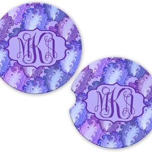 May include: Two round car coasters with a purple feather pattern and a monogram in a light purple frame. The coasters are designed to fit in a car's cup holder and protect the interior from spills.