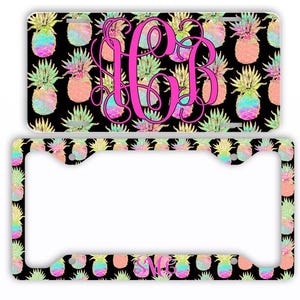 May include: A license plate and frame set featuring a black background with a repeating pattern of colorful pineapples. A large pink monogram is centered on the license plate. The frame also has a pineapple pattern.